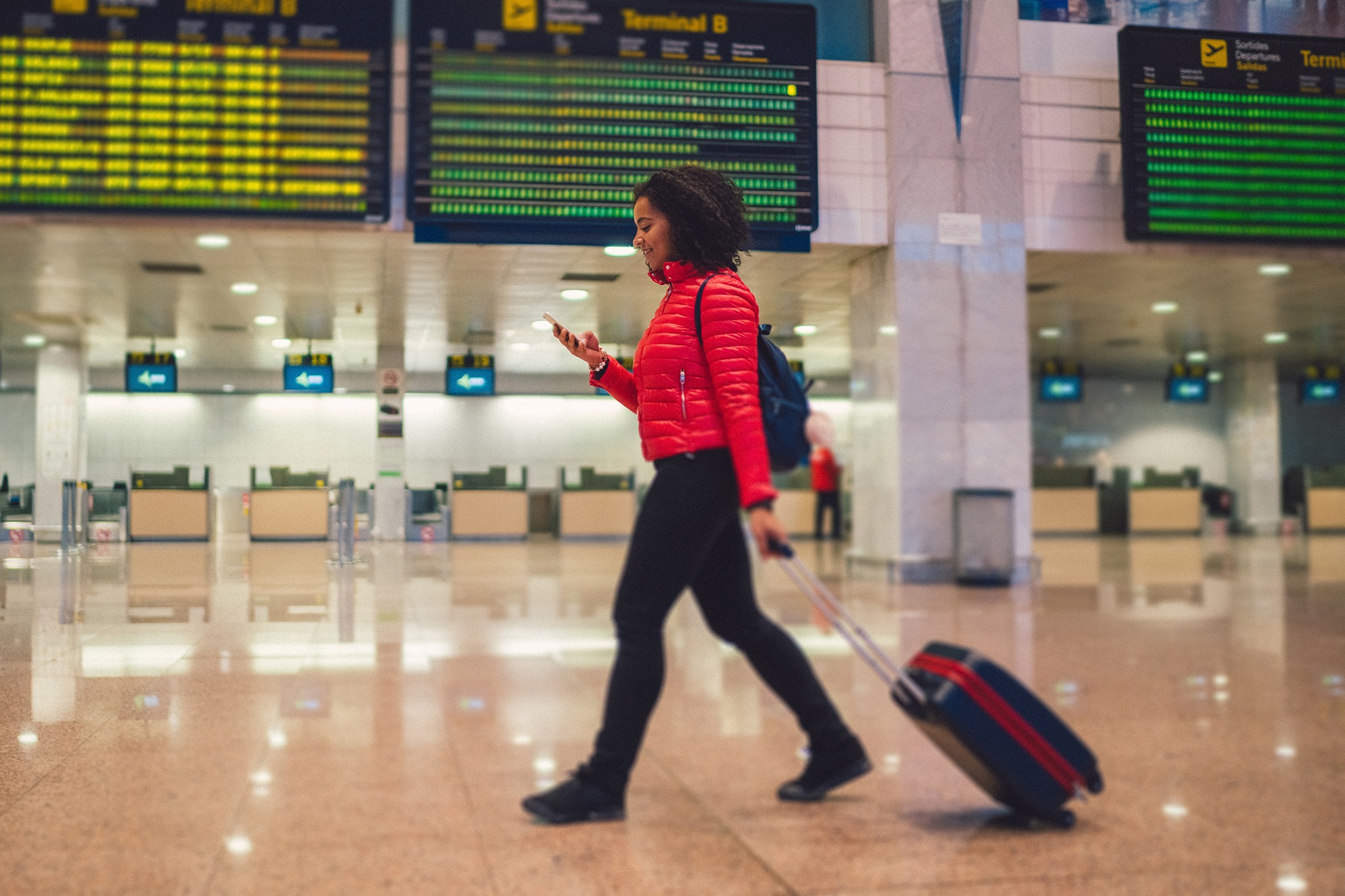 Young woman with wheeled luggage walking at the airport and texting